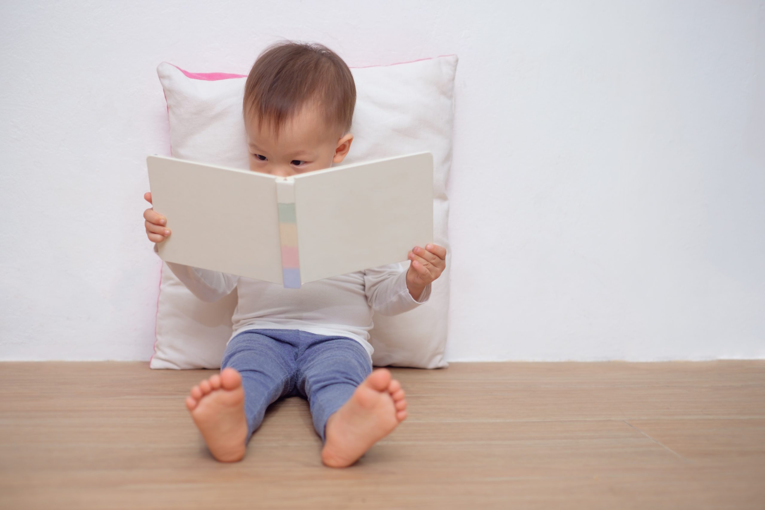 Cute little Asian 18 months / 1 year old toddler boy child sitting on floor, leaning against pillow, looking at a book near white wall, Language development, Improve concentration & focus concept
